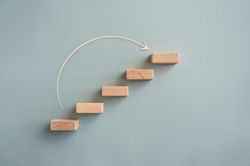 Wooden blocks forming ascending steps on a light-blue background, with an arrow illustrating upward movement, symbolizing growth, progress, or achievement