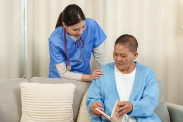 Obraz premium Young asian female physical therapist gently smiling placing hand on elderly female patient's shoulder while standing beside sofa during home rehabilitation supporting recovery encouragement
