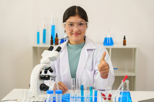 Caucasian teenage female student wearing lab coat gloves goggles giving thumbs up smiling at camera sitting in science classroom with laboratory glassware microscope indicating success in experiment