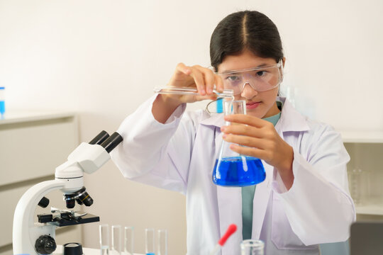 Caucasian teenage schoolgirl wearing lab coat and protective goggles pouring blue chemical liquid from test tube into flask during science class experiment in laboratory with microscope on table