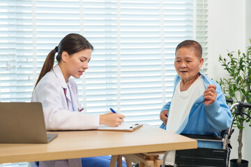 Obraz premium Asian senior woman sitting in wheelchair lifting cardigan during medical consultation with female doctor writing on clipboard, showing health checkup interaction and elderly patient care in hospital.