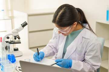 Caucasian high school girl scientist wearing gloves lab coat goggles writing on clipboard while conducting science experiment in chemistry classroom with test tubes beakers and microscope on table