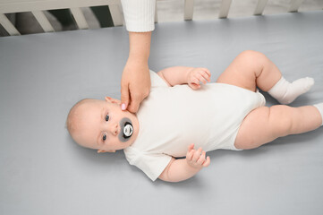 Cute baby with pacifier lying in crib, mother taking care of infant caressing him, tickling his...