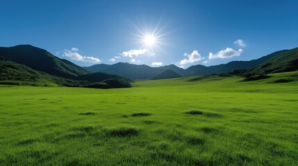 Fototapeta premium Bright sun shining over a vast green meadow surrounded by distant mountain ranges under a clear blue sky.