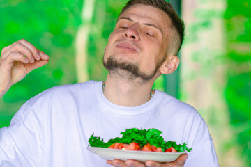 Smiling bearded man in a white t-shirt holding a plate of fresh vegetable salad with lettuce and tomatoes, on a bright green natural background.


