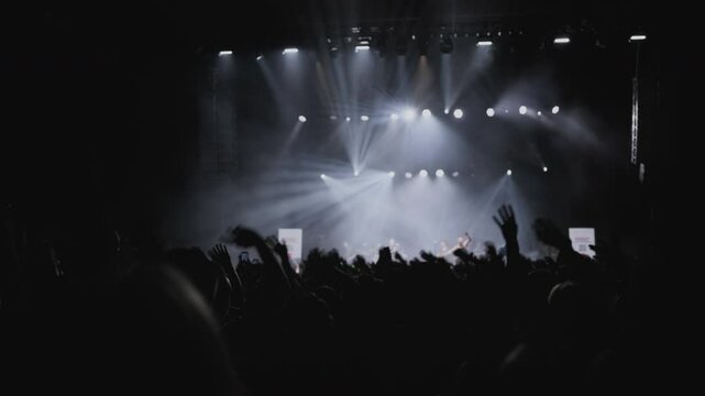 Concerthall with light show.Fans with rased your hands and wave for music against background white, magenta and blue spotlight on stage. Medium close-up shot. Slow motion. 