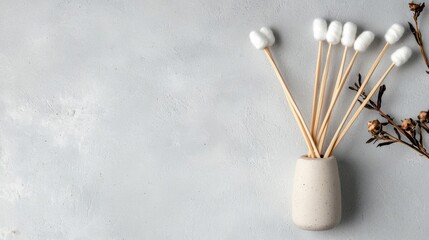 Bunch of cotton buds in a white ceramic vase. the vase is placed on a light grey textured background. the cotton buds are arranged in a bunch with wooden sticks sticking out of the top.