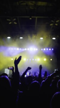 Music light show. Dark silhouette of raised hands of fans in wave motion. Blue and ellow background from stage. Medium close-up shot.