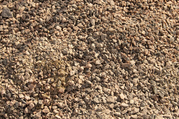A large pile of gravel rests on the ground at a construction site. Sunlight illuminates the textured surface, showcasing various shades of brown and gray stones