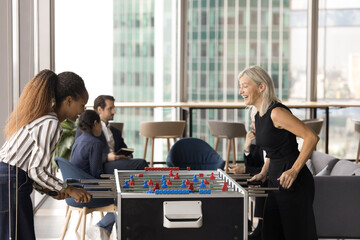 Relax from job. Two female colleagues of diverse age ethnicity business partners managers aged team leader young worker enjoy playing kicker game at modern office space while teammates talking at desk