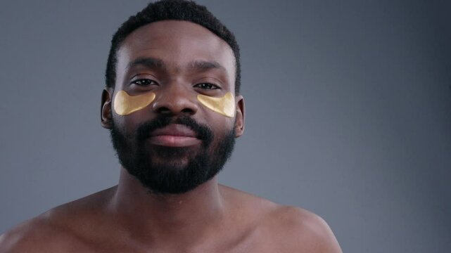Attractive african young man with under eye golden patches caring for his facial skin looking straight at camera on grey background.
