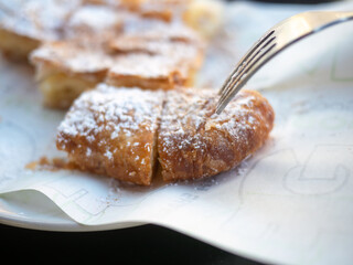 Closeup of hands eating traditional Greek bougatsa, a sweet cream pie pastry with filo dough, dusted with powdered sugar and cinnamon. Popular breakfast or snack from Thessaloniki and Serres, Greece.