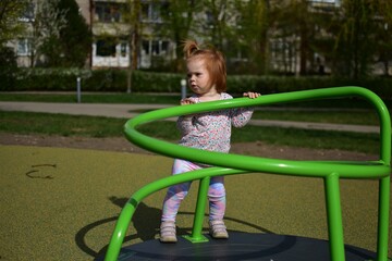 Fototapeta premium Baby girl standing on spinning equipment in children playground in city park in warm weather spring. Toddler girl having fun spinning around in public playground in sunny day spring.