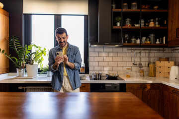Happy man enjoying his smartphone while preparing a delicious meal in a modern kitchen, surrounded...