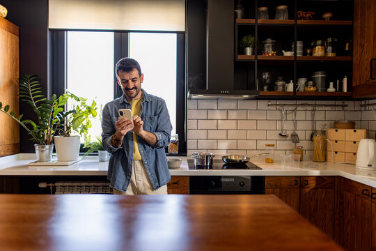 Happy man enjoying his smartphone while preparing a delicious meal in a modern kitchen, surrounded by plants and a bright window