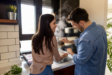 Happy couple cooking together in a modern kitchen, enjoying the warmth and steam rising from a pot...