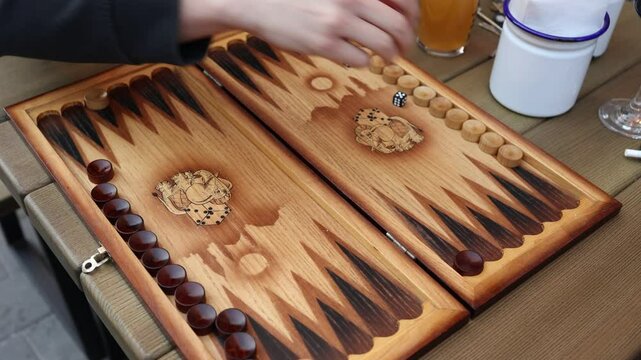Hands of two players in the middle of a backgammon game, played on a decorative wooden board at a cafe table.
