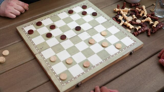 A casual checkers game is being played on a wooden board outdoors, with chess pieces scattered nearby and hands visible.