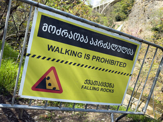 Multilingual warning sign near waterfall in Georgia prohibits walking due to rockfall danger in...