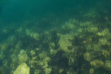 A top-down view shows clear, dark turquoise water with sunlight dappling the submerged, moss-covered rocks on the seabed near the coast