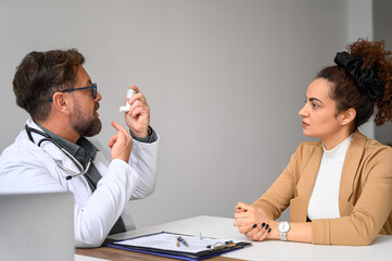 Male doctor in lab coat showing and explaining how to use asthma inhaler to young woman in hospital