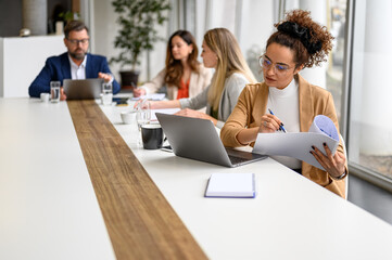 Confident businesswoman reviewing documents and working on a laptop while colleagues collaborate in...