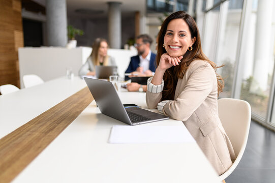 Confident businesswoman with laptop on table smiling at camera while legal colleagues collaborating in the background