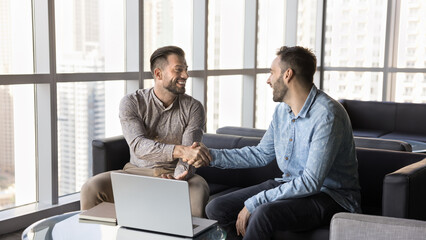 Two young smiling businessmen seated in modern office lounge, shaking hands, gesture of successful...