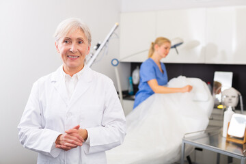 Obraz premium Portrait of smiling confident professional elderly woman cosmetologist wearing white uniform standing in modern medical aesthetic office..
