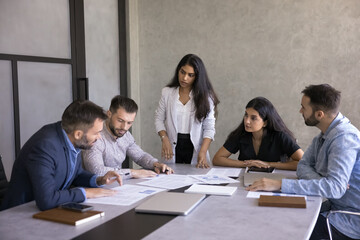 Group of five professionals engaged in brainstorming or decision-making, take part in collaborative meeting seated around conference table, discuss documents, charts, and report, review paperwork data