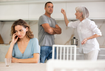 Fototapeta premium Young girl is sitting at table in kitchen quarreling with her mother-in-law and husband
