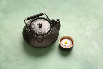 Teapot with cup of natural chamomile tea and flower on green grunge background