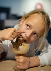Blonde girl greedily eating ice cream with a slice of Borodino rye bread, holding a glass goblet, sitting at a table in a restaurant, looking away