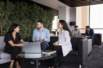 Three teammates lead talk seated in office lounge area, engaged in focused conversation,...