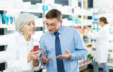 Obraz premium In pharmacy, senior woman apothecary holds package with therapeutic cream for sensitive skin in hands and talks with male client about nuances of care.