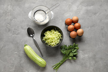 Bowl with grated zucchini and ingredients for preparing fritters on grey background