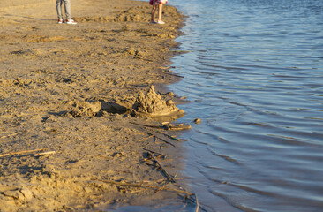 The shore of a lake with wet sand and traces of children's play. Unfinished sand structures and the feet of children standing by the water. The scene is captured in the warm light of the afternoon sun