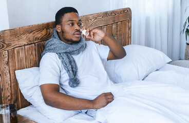 Cold And Flu Treatment. Sick African American Man Using Nasal Spray Having Rhinitis And Sinusitis Sitting In Bed In Bedroom At Home. Nasal Stuffiness And Runny Nose Health Problem, Healthcare Concept