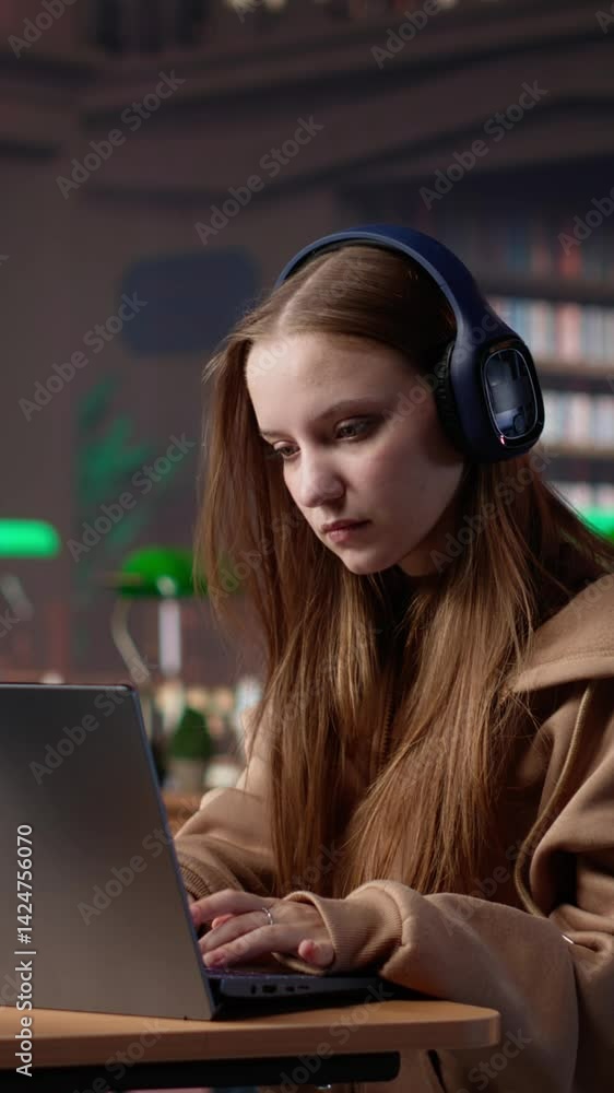 Poster Vertical Video Young student attending to a web lecture with headset in the campus library, takes notes and learns about politics in an online meeting. Focused pupil during class lesson. Camera A.
