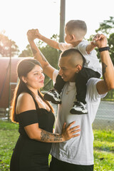 young latin couple in the park walking with their child, man carrying the child on his shoulders while the woman looks at her husband, sunset in background