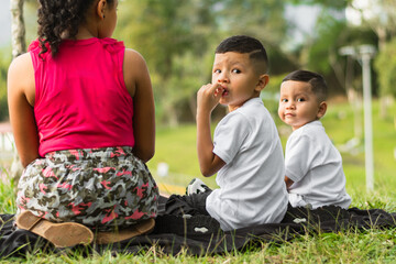 view of the back of two boys and a girl sitting in the park, Latino children looking back, turning around to look