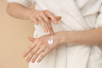 Woman in white bathrobe applying cream onto hands on color background, closeup