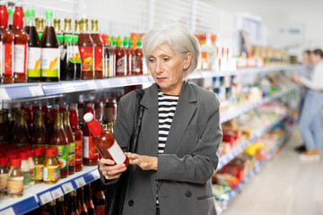 Focused aged woman reading labels on bottles with sauces in supermarket, carefully examining ingredients and expiration date while shopping for groceries