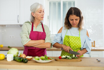 Elderly mother teaching adult daughter how to cook vegetable salad in the kitchen