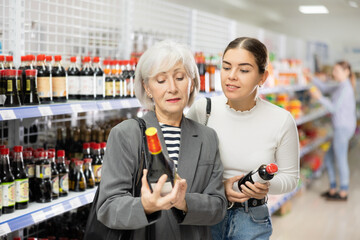 Interested elderly woman discussing choice of soy sauce with young girl while standing by shelves...
