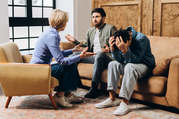 Teenage boy holding his head and father talking to female psychologist