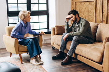Tired adult man sitting on a couch and talking to female psychotherapist