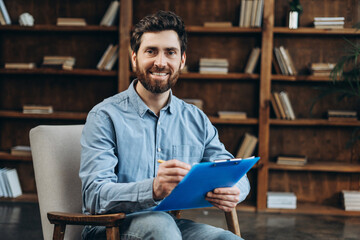 Smiling male therapist writing notes sitting on armchair posing at camera in the office