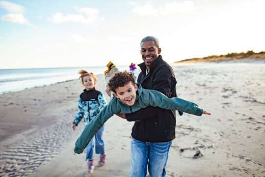 Happy African American dad playing airplane game with kids on beach during family vacation
