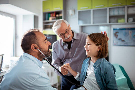 Smiling doctor using stethoscope to check young girl at medical clinic with grandfather nearby